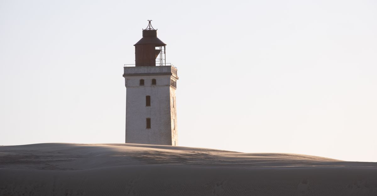 A lone lighthouse standing steady above sand dunes in soft evening light.