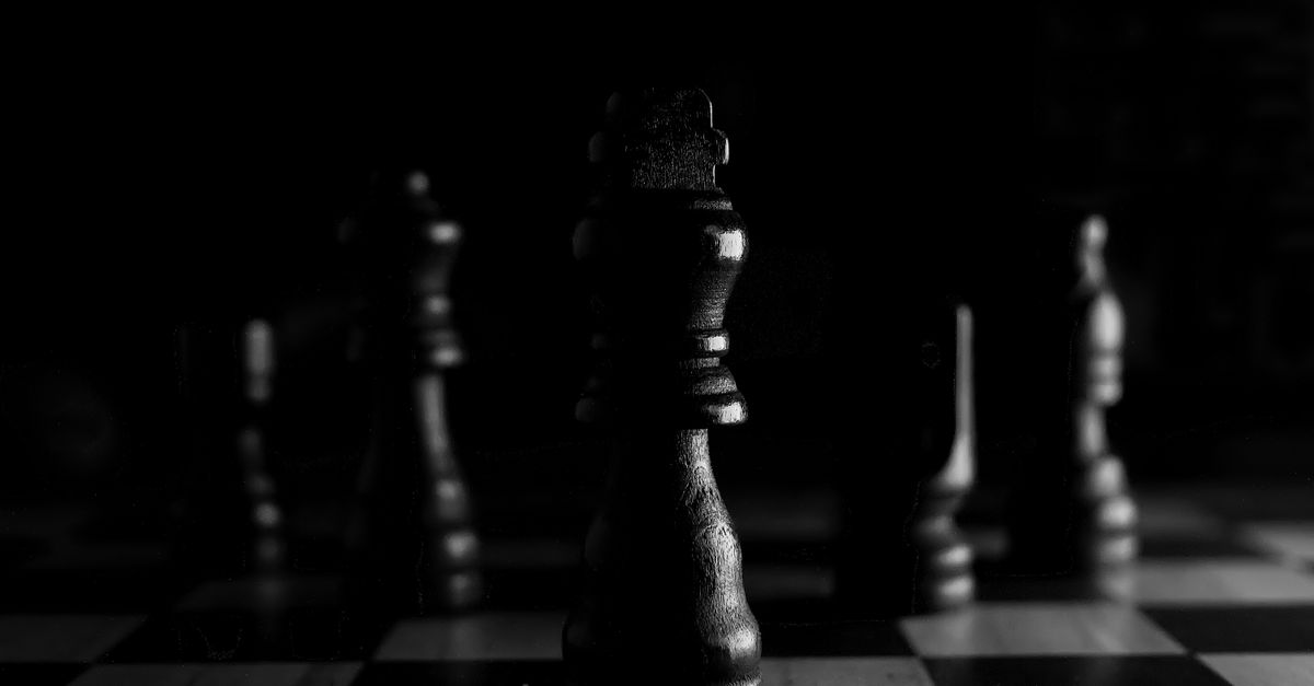 A single chess piece lit in the dark on a checkered board, other pieces waiting in shadow